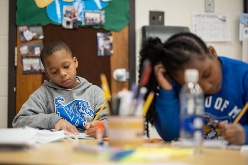 Two young students sit at their desks concentrating on their schoolwork while wearing blue, white and grey sweaters.