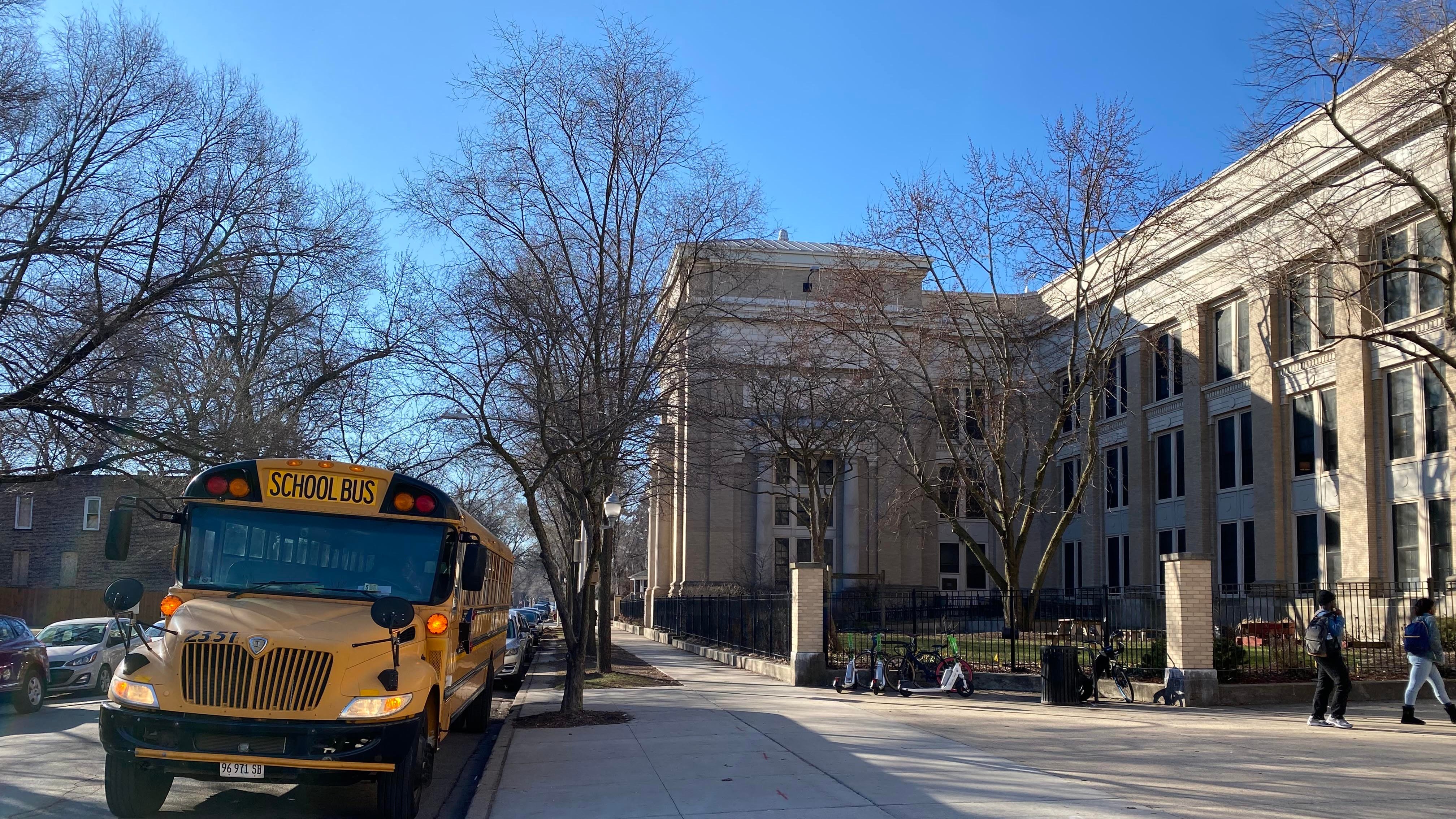 A yellow school bus is parked outside of a large stone building with a large sidewalk in the foreground and blue sky in the background.