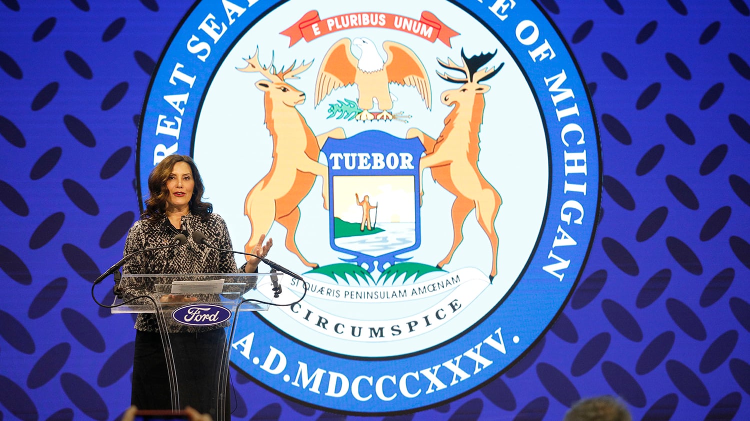 A woman wearing a dark top and a black skirt stands at a podium in front of a large Michigan state seal.