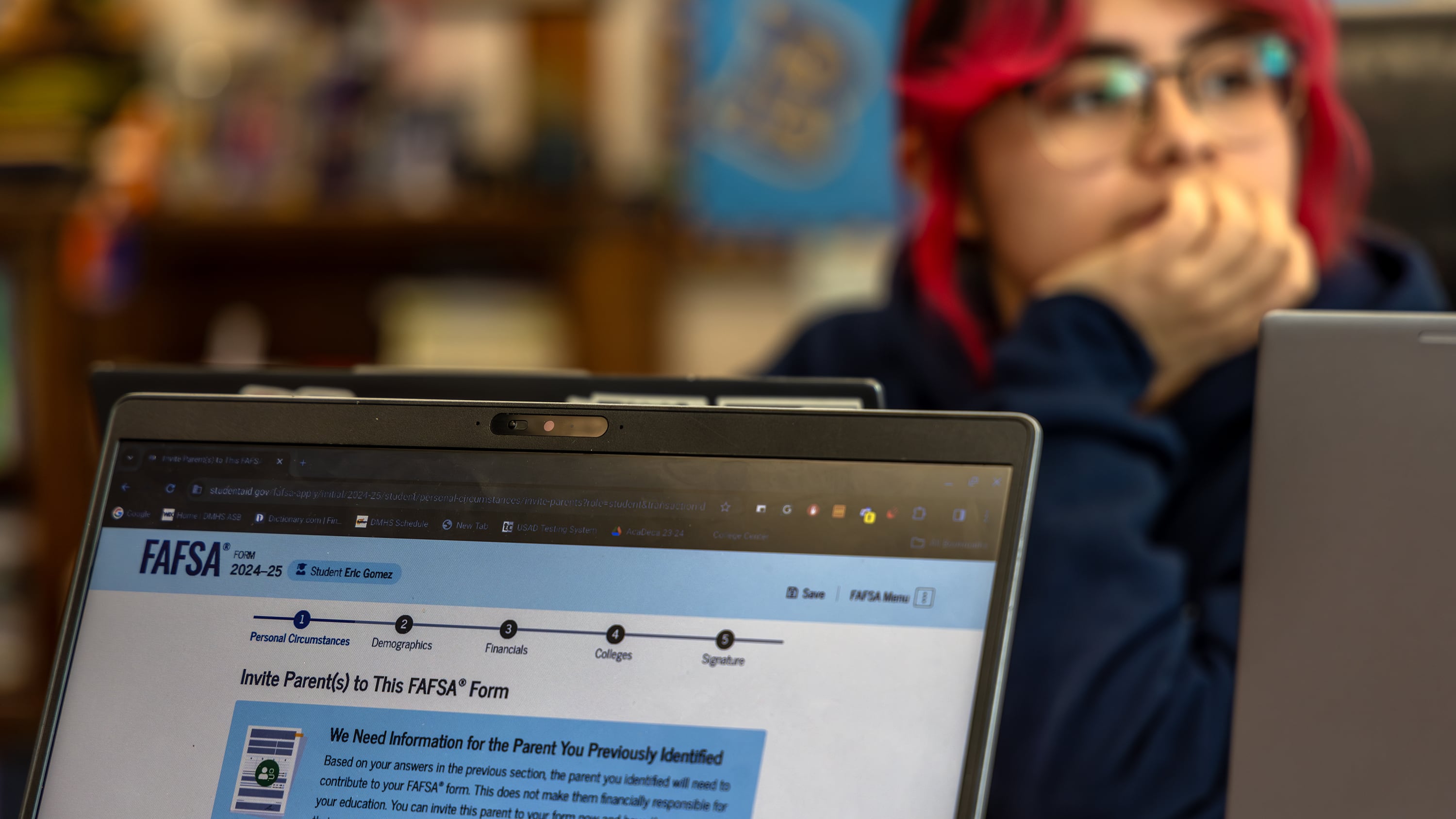 The top half of a laptop screen is in focus with the word "FAFSA" in view with a student wearing glasses and has red and black hair in the background.