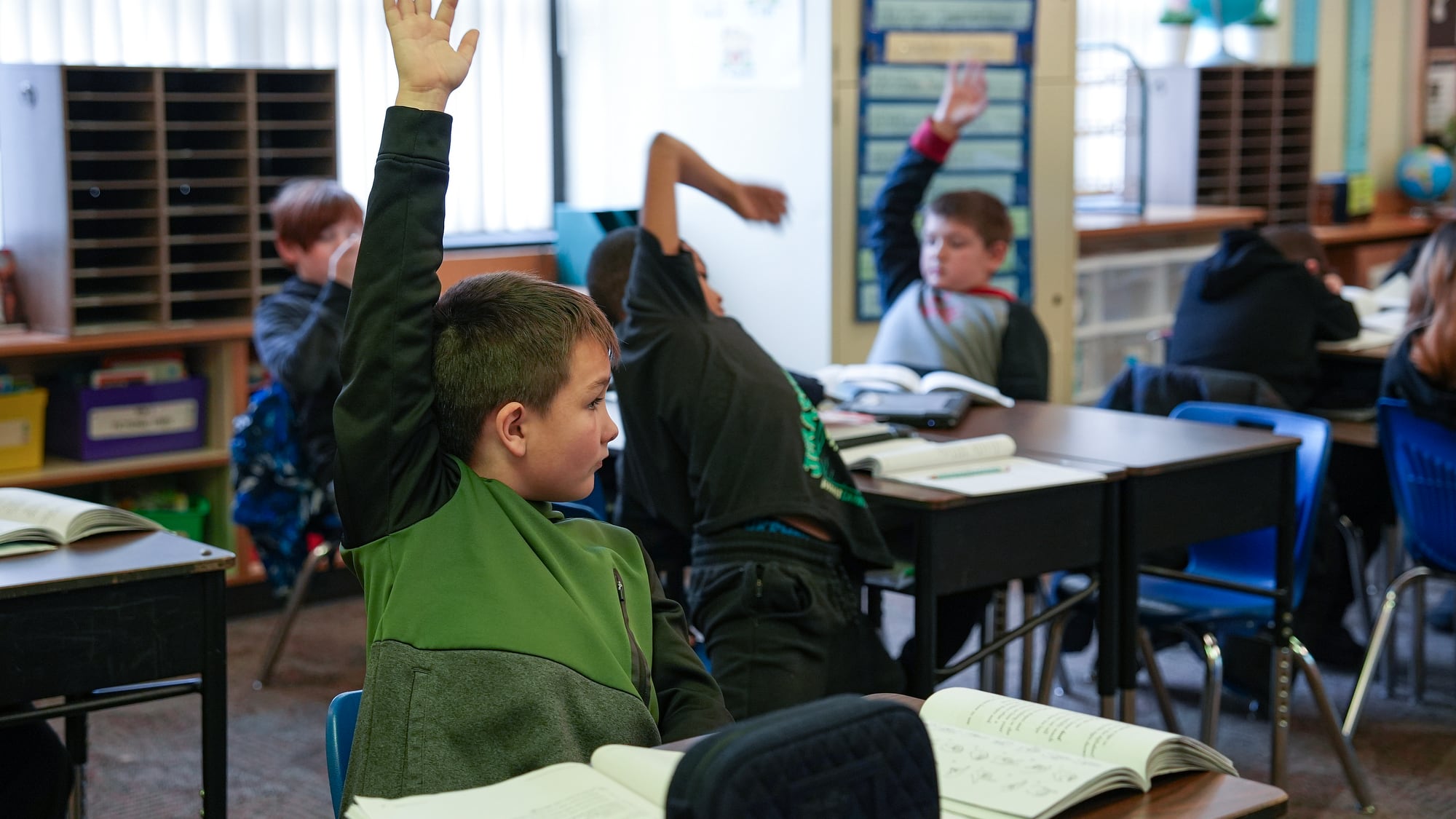 A group of young students sitting at their desks raise their hands in a classroom.