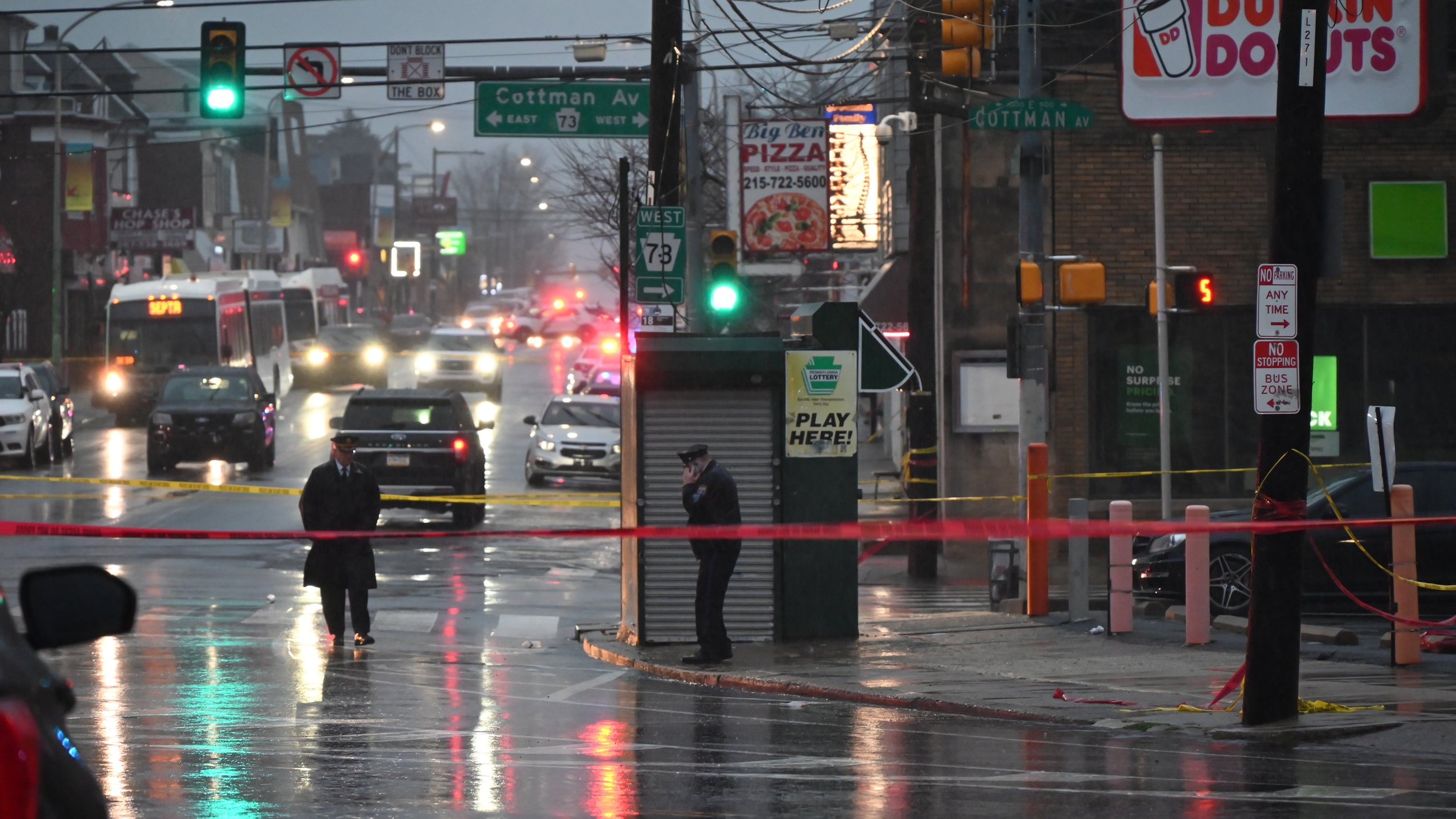 Two police officers stand on the sidewalk and in the street with a red caution tape across the street with cars in the background.