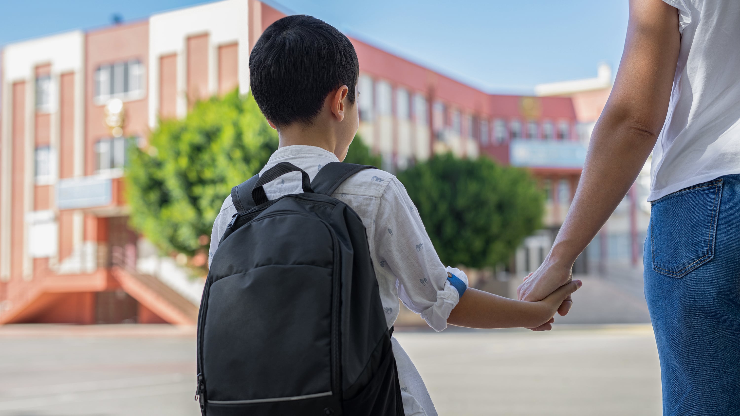 A young student wearing a black backpack holds their parent's hand with a school building in the background.