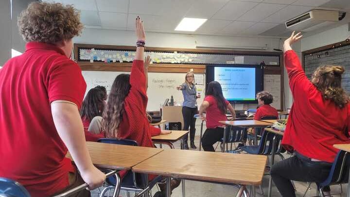 A teacher at the front of a classroom points to students in red shirts who have their hands raised to volunteer for an assignment.