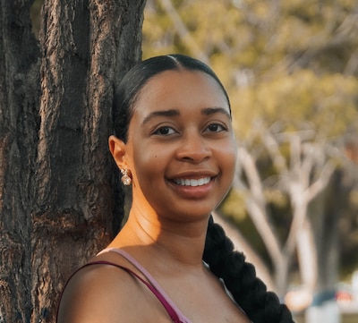 Headshot of a Black woman with long braided hair. She leans against a tree.