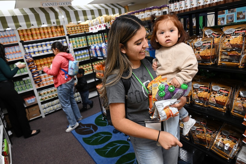A woman carries her daughter and a handful of packaged foods with a couple of people shopping in the background. There are shelves full of food surrounding them.