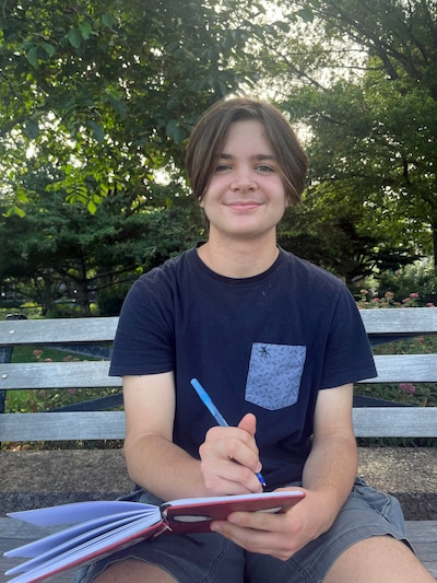A young person wearing a dark blue shirt and holding a pen and notebook sits on a wooden bench outside with green trees in the background.