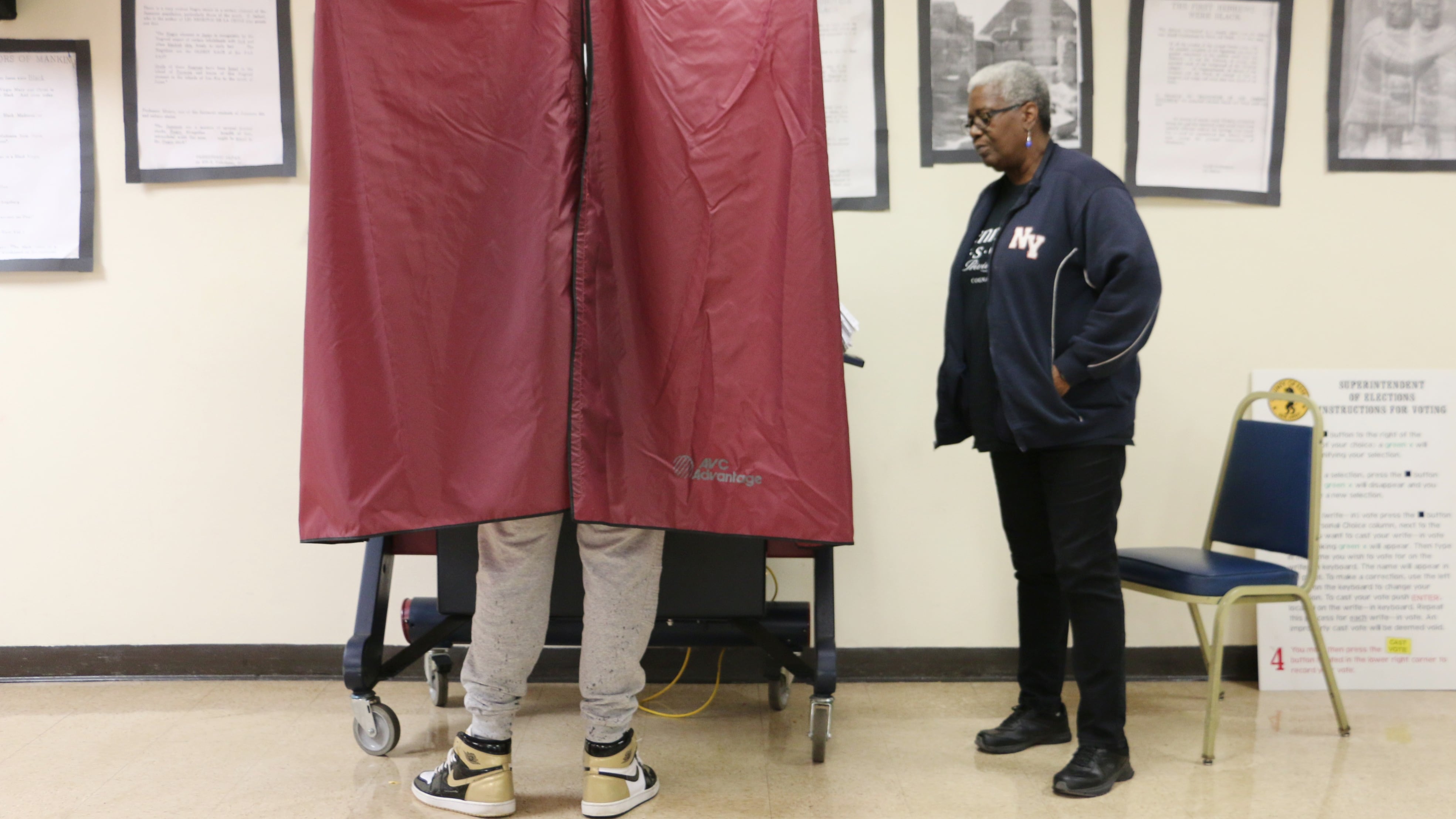 One person stands outside of a voting booth while one person is using the voting booth in a room with large picture frames on the wall in the background.